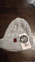 Light gray knit Buff reversible hat laid flat showing cable knit pattern and ribbed cuff with leather logo patch and original tags.