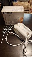 Philips hand mixer with two metal beaters placed next to its original packaging box on a wooden surface.
