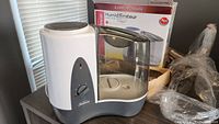 White and gray Sunbeam humidifier with cracked transparent water tank on tabletop, original box in background
