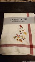 Photo of folded off-white tablecloth with red border stripes and autumn leaves embroidery, with packaging label 'TABLECLOTH set the perfect table!'