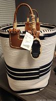 Front and side view of the large woven bag with white and navy stripes, brown leather handles wrapped with beige cord, attached brand tags visible.