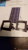 Photo showing dual 60 LED floodlight heads with black aluminum casing next to solar panel with blue photovoltaic cells