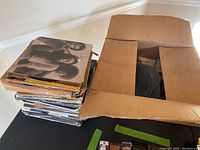 Photo of a stack of vinyl record albums and 45 RPM records alongside a cardboard box containing loose records, showing overall quantity and condition.