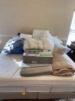 Photo of assorted bedding items arranged on a bed including white and blue pillows, beige and striped duvets, and a mattress fitted with a white sheet.