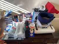 Lot of health and personal care items on a wooden table including foot massager, neck massager, blood pressure cuff, razors, toothbrushes, wrapping tape, ice bags, and a TENS 3000 device.