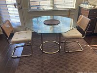 Octagonal glass top table with two brass frame chairs with upholstered seats and rattan backs positioned on patterned carpet near window; shows overall set from an angle.