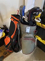 Photo shows two golf bags placed side by side in garage area, one grey and brown Callaway Golf bag, the other orange and black Adams Golf bag, both filled with golf clubs.