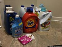 Image showing multiple bottles of laundry supplies arranged on the floor including Tide orange liquid detergent, white Shout stain remover, blue Fantastik cleaner, Clorox bleach, and other bottles.