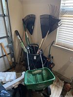 Wide view showing collection of leaf rakes, shovel, pruning clippers, pickaxe stored in black plastic container and Scotts broadcast spreader in front.