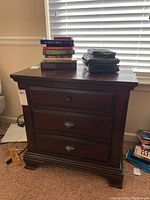 Front view of dark cherry wood nightstand with three drawers, showing books and small items on top.
