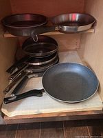 Photo showing multiple cake pans, skillets, sauce pots with glass lids stacked inside cabinet