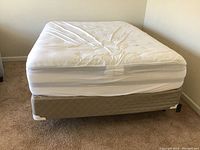 Full view of queen size mattress with fitted white sheet and beige box spring on carpeted floor.