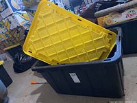 Five large black plastic storage totes each with a yellow lid. Lids have reinforcement ribs. Totes are empty, placed in garage area with other items visible in background.