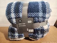 View of folded blue and gray plaid sherpa throw blanket with attached product label and gray strap.