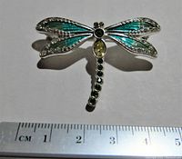 Photo of silver dragonfly brooch pin with blue-green enamel wings and round green and yellow gemstones, approximately 4 cm wide, shown with ruler for scale.