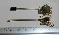 Two vintage stick pins with green jade stones and gold-tone metal; one oval floral design, one butterfly filigree design, shown with ruler for size reference.
