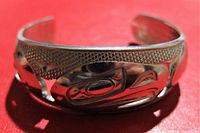Front view of the silver cuff bracelet showing the polished carved West Coast Native art animal face design against textured background.
