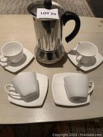 Top view of 4 white espresso cups on square saucers arranged around a metal stovetop percolator with black handle and knob on a wooden table.