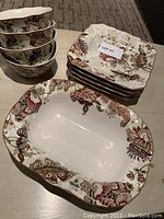 Stack of 4 soup bowls, stack of 4 dessert plates, and 1 serving bowl displayed on a table, all with a floral pattern in burgundy, beige, and green.