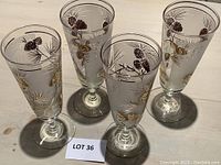 Four clear parfait glasses with gold leaf decoration of pinecones and pine needles arranged on a table with lot sign visible.