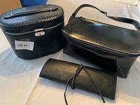 Three black makeup cases: one textured hard case with handle and lock, one rectangular soft case with shoulder strap, and one folded brush carrier.