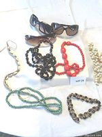 Photo showing 5 costume jewellery necklaces and 3 pairs of sunglasses arranged on a white surface with lot number.