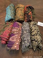 Seven rolled and folded pashmina scarfs/shawls in varied colors and patterns arranged on a wood surface to display diversity of styles in the lot.