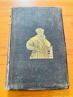Front cover of the book showing black textured cloth with a gold embossed image of a bearded man in historical dress.