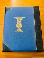 Closed cover of the 1957 Haggadah book showing blue cloth with black leather corners and spine, featuring a gold embossed urn design in the center.