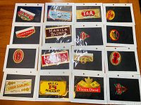 Full overhead view of fifteen cigarette labels individually mounted in sleeves on black backing cards, arranged in a 4x4 grid with one empty slot