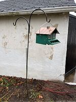 View of the bird feeder hanging from the metal stand outdoors, showing overall setup and contrast with a white building wall.