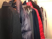 Photo showing several jackets and coats including black wool coat, black and red puffer jackets, grey sweater with fur collar, and others hanging on rack.