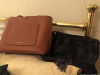 Photo showing both a brown leather tote with front pocket and gold logo and a black large purse with tassels on a bed.