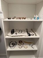 Wide view of three white shelves holding assorted costume jewelry including brooches, necklaces, bracelets, and watches.