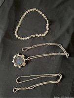 Three silver-tone necklaces displayed on black background. One with linked floral square motifs, one chain with round pendant, and one simpler chain.