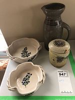Four items placed on a table against a wall: two ceramic serving dishes with blue floral designs, rustic jam or honey pot with lid and spoon, and a dark pottery pitcher with handle.
