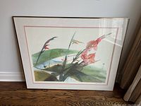 Framed floral artwork resting against a white wall on a wood floor, showing signature area and floral details.