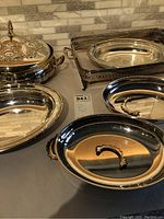 Photo showing seven silver plated serving dishes of various shapes on countertop, including an oval dish with lid, rectangular tray with pierced edges, and two dishes with broken handles and detached handles included.