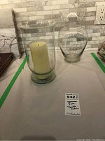 View of the glass hurricane candle holder with white candle inside next to the clear glass vase on a white surface with green tape.