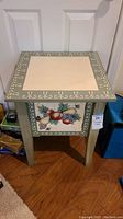 Full view of square wooden accent table with handpainted fruit design on side panel and cream-colored top inset