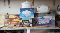 Three boxed stoneware and porcelain bake and serve dishes including Bobby Flay gratin dishes, porcelain casserole with lid and stand, and large stoneware baker with stand.