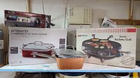Overall view of the three main items including boxed Ambiano slow cooker, boxed Swissmar SwissRaclette grill, and Copper Chef pan with glass lid.