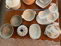 Overview of lot on wooden surface showing white Corning Ware pots, porcelain container with fruit motif lid, Galenn pottery bowls, and white ceramic bowls and tray.