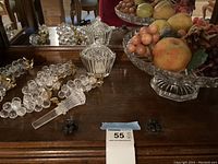 Photo showing two ribbed glass or crystal candy dishes with lids, clear and reflective, and one glass pedestal fruit dish filled with artificial fruit on a wooden surface.