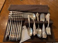 Overview of the full cutlery assortment including knives, spoons, forks, and serving spoons arranged on a dark cloth holder.