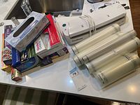 Photo showing FoodSaver vacuum sealer unit with five vacuum sealer rolls stacked beside it on a white surface near a kitchen sink.