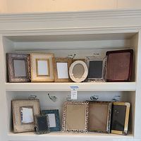 View of two shelves containing 11 assorted picture frames of different shapes, sizes, and decorative styles.