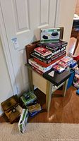 Photo showing stacked board games including Rummy Jack, Chinese Checkers, Monopoly, Tri-Ominos, along with card decks and holders on small table.