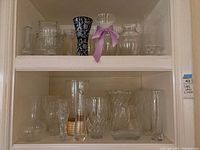 Two shelves filled with various glass vases of different shapes, sizes, and designs, some clear glass, one dark-colored, and one with a ribbon.