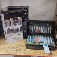 Two MAXIM Circleware 6-piece stemware sets in boxes and two Versailles 24kt gold decorated crystal glasses boxes displayed on plywood shelf.